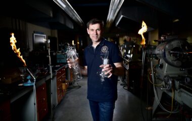 Mark Toonen holding blown glassware in a glass blowing shop with flames and machinery in the background