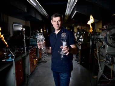 Mark Toonen holding blown glassware in a glass blowing shop with flames and machinery in the background